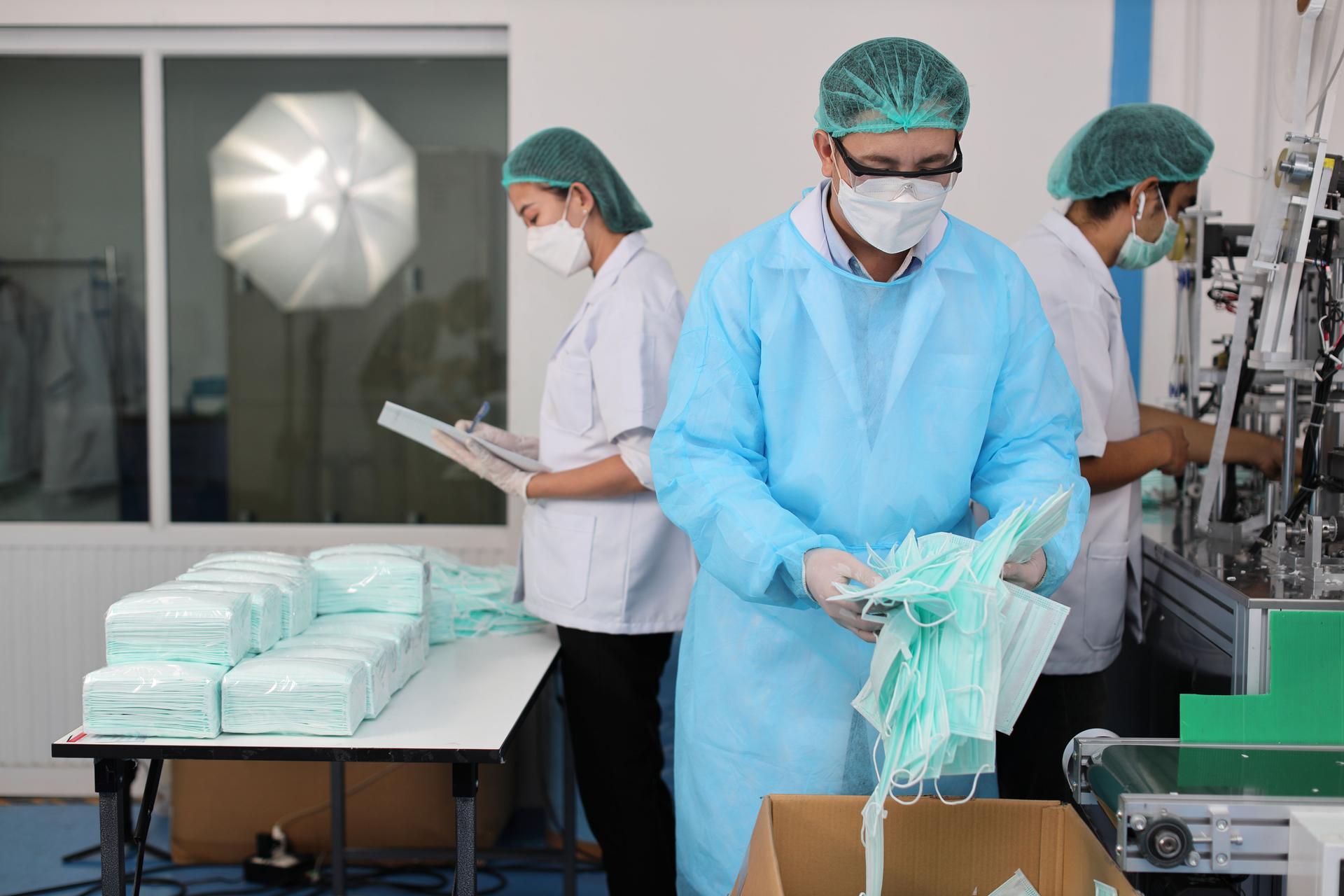 Worker man and woman in personal protective equipment or PPE inspecting checking quality of mask and medical face mask production line in factory, manufacturing industry and factory concept.